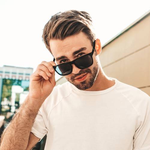 Portrait of handsome smiling stylish hipster lambersexual model.Man dressed in white T-shirt. Fashion male posing in the street in sunglasses